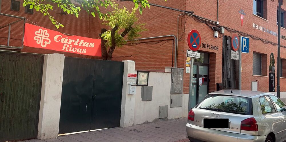 Entrada de Cáritas Rivas: portón verde con el rótulo de Cáritas Rivas, junto a la Biblioteca Almudena Grandes, en la calle del Grupo Escolar